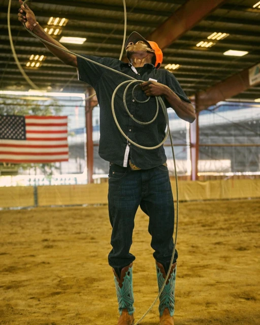 A man in jeans and cowboy boots throws a lasso high in the air during a rodeo performance indoors, with the American flag in the background.