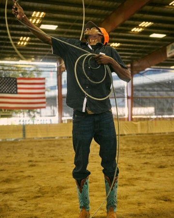 A man in jeans and cowboy boots throws a lasso high in the air during a rodeo performance indoors, with the American flag in the background.