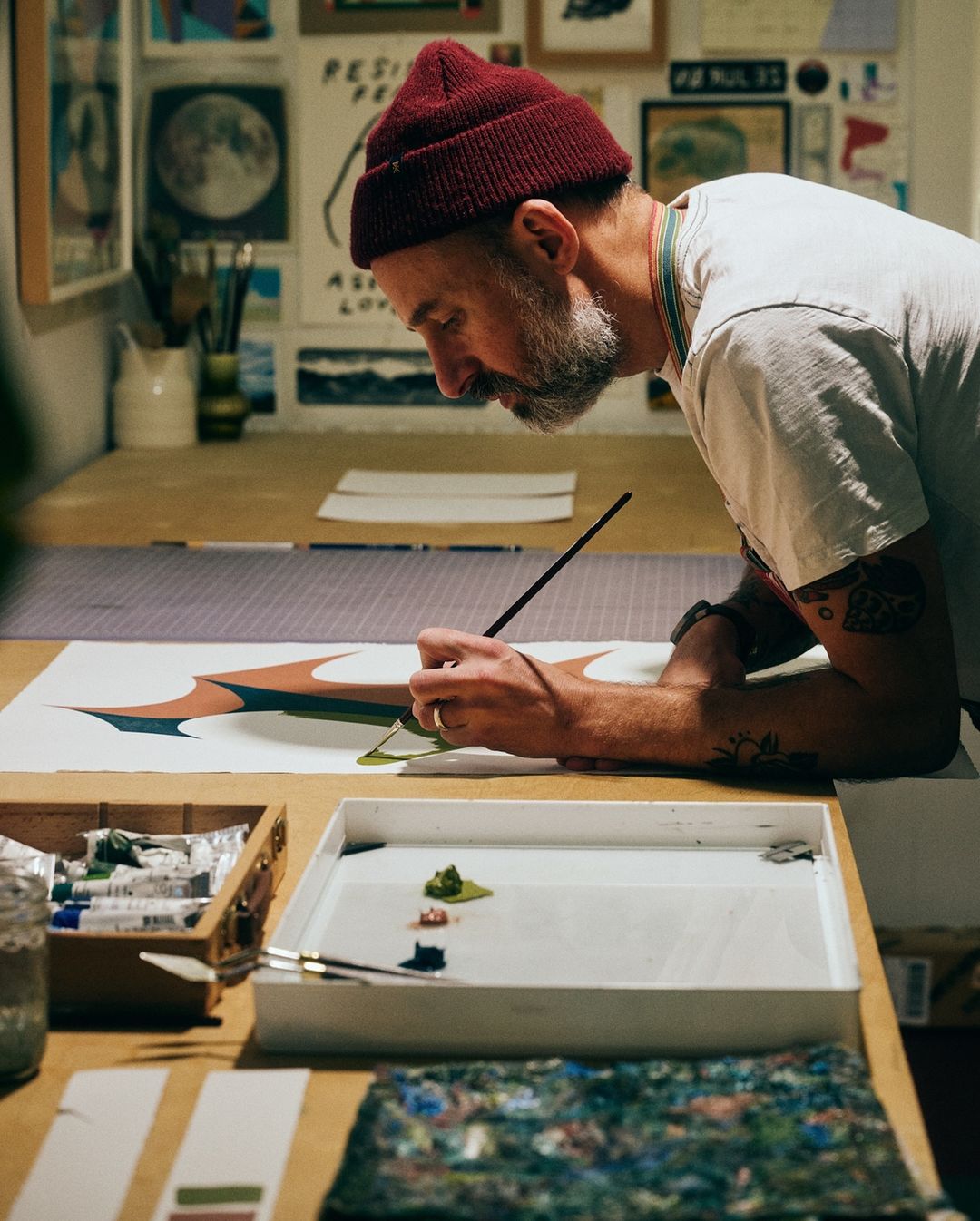 Artist with beard and red beanie at work painting on canvas in a studio setting; creative process captured in natural light.