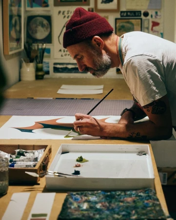 Artist with beard and red beanie at work painting on canvas in a studio setting; creative process captured in natural light.
