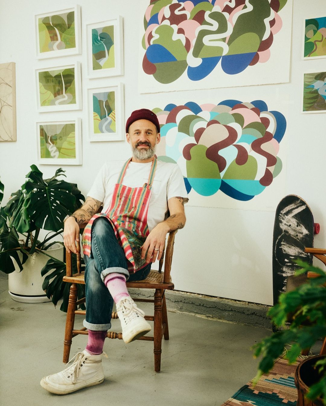 Artist sitting on wooden chair in art studio with paintings on the wall, wearing a red beanie, striped apron, and white high-top sneakers.