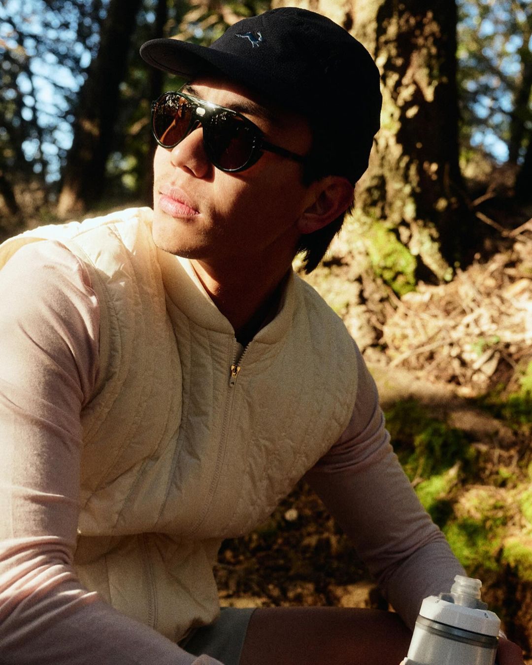 Outdoor portrait of a young adult man wearing a hat and sunglasses in a forest setting with a bottle of water.