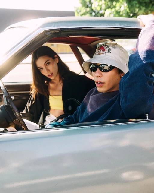 A young adult man and woman sit inside of a light-colored vintage car with a blue exterior under bright sunlight outdoors.