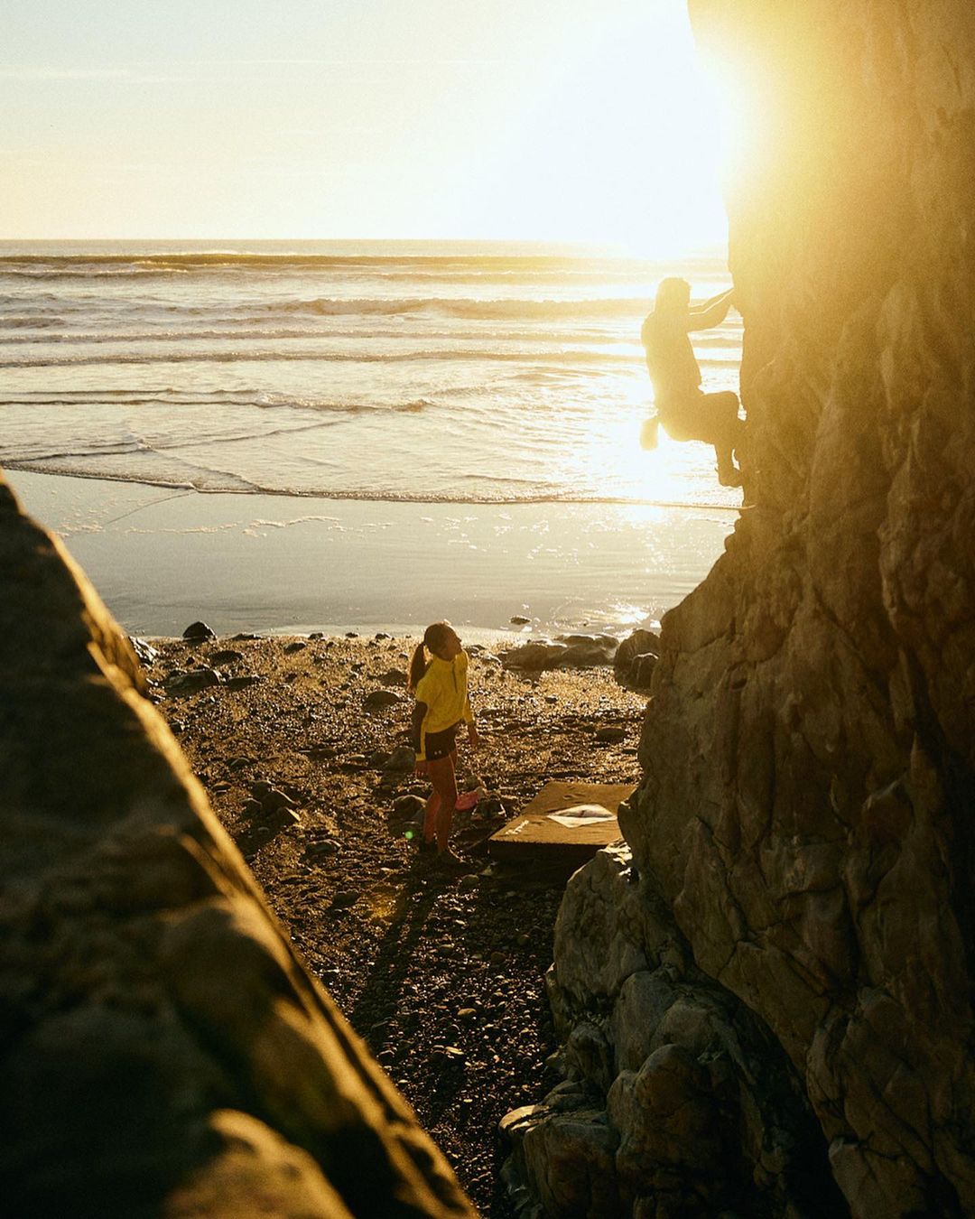 Rock climber ascending cliffside at sunset, with child on beach below. Ocean waves visible in the background. Warm, golden light.