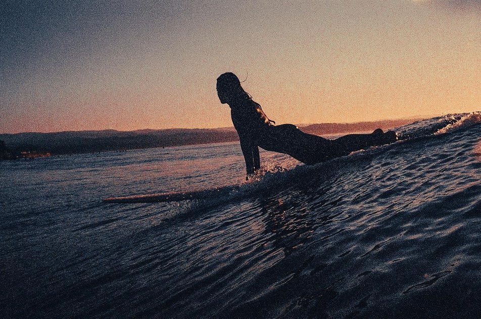 Silhouette of a surfer in the ocean at sunset, paddling on a surfboard towards the horizon with distant mountains in the background.