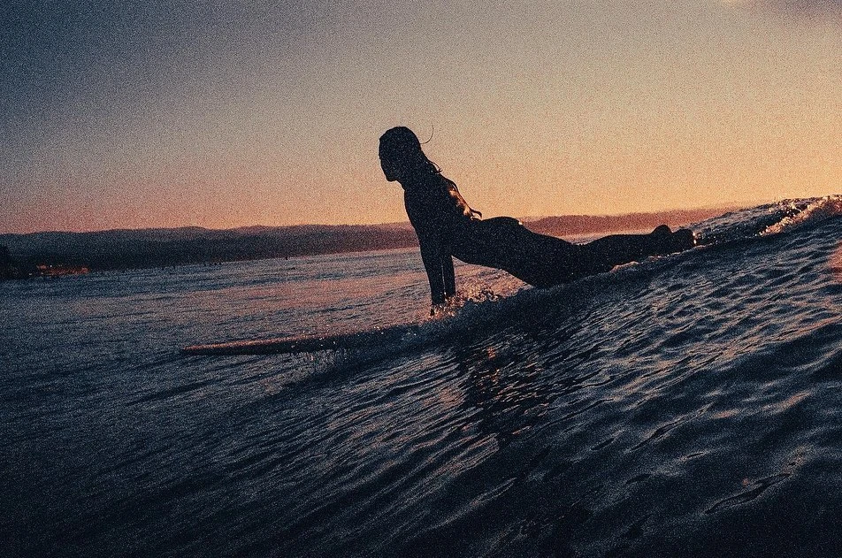 Silhouette of a surfer in the ocean at sunset, paddling on a surfboard towards the horizon with distant mountains in the background.