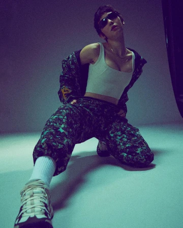 Model wearing dark sunglasses, white tank top, and patterned tracksuit kneeling against a plain backdrop.
