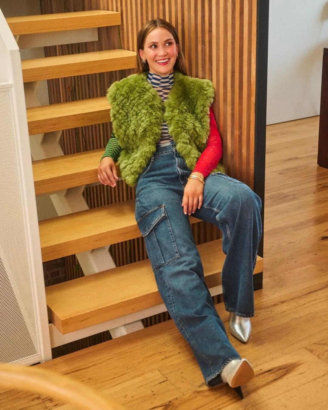 A woman is sitting on wooden stairs wearing a colorful striped top with a green faux fur vest, wide-legged jeans, and metallic silver boots.