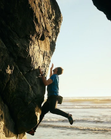 Man in blue shirt climbs a large rock formation on a beach near ocean waves; wearing climbing shoes and gear with clear blue sky.