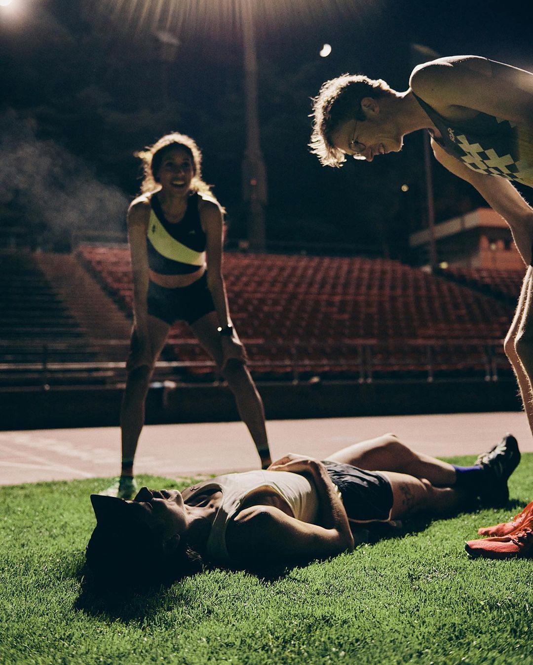 Track and field athletes pause after a race, one lying on the grass, two standing looking down in an outdoor stadium at night.