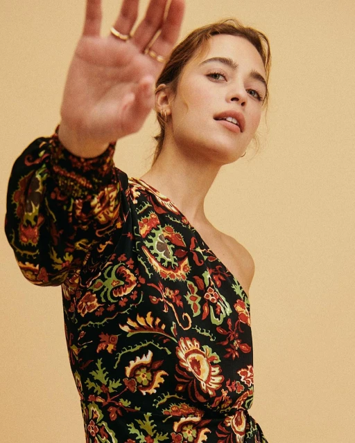 Attractive brunette woman in an elegant, one-shoulder floral dress poses in a studio with her hand outstretched toward the viewer.