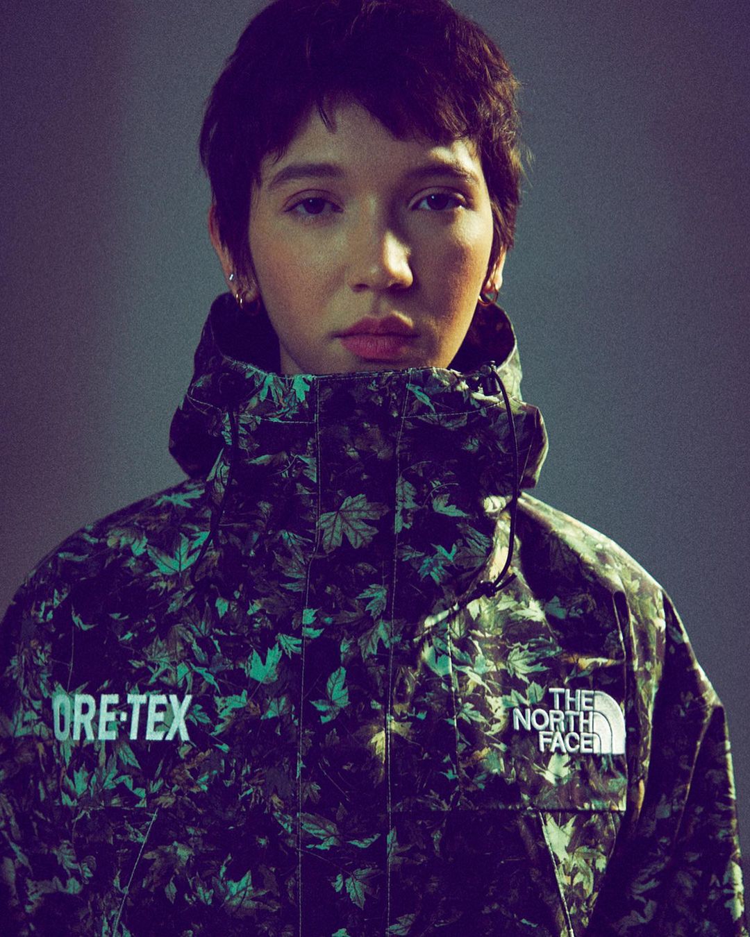 A woman with short dark hair and light skin wears a leafy green-and-black jacket with visible brand logos against a grey background.