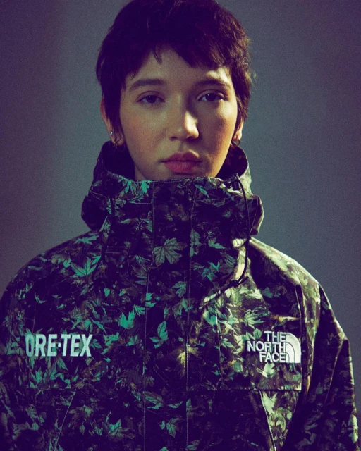 A woman with short dark hair and light skin wears a leafy green-and-black jacket with visible brand logos against a grey background.