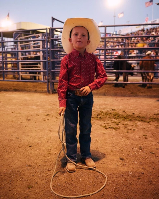 Young boy in cowboy attire stands in a dirt arena with a cowboy hat, red patterned shirt, jeans, and boots, holding a lasso rope.