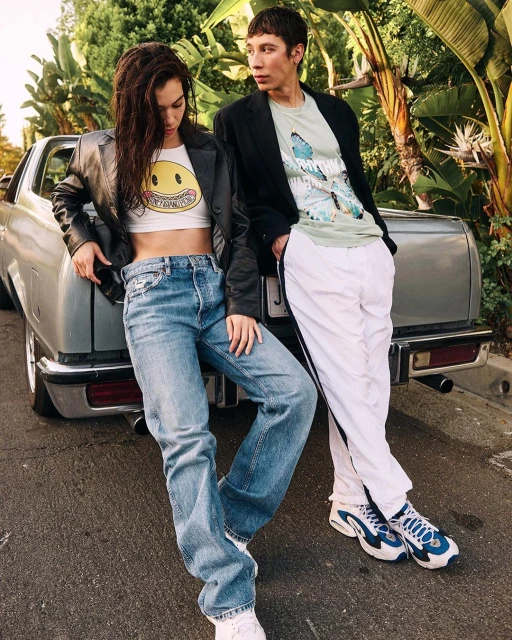 Two people posing outdoors next to a grey car; a woman in jeans and a crop top with a smiley face and a man in white pants.