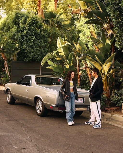 A young couple stands next to a silver El Camino on a tree-lined street, with lush greenery creating a natural backdrop.
