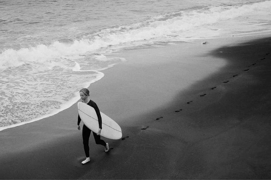 Monochrome photo of a surfer walking on a beach with a surfboard, footprints visible in the sand and waves in the background.