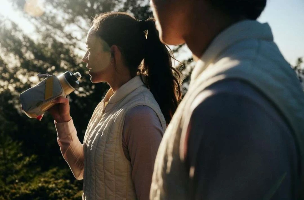 Woman drinking from water bottle outdoors near another person. Healthy hydration, fitness activity in a sunny, natural environment.