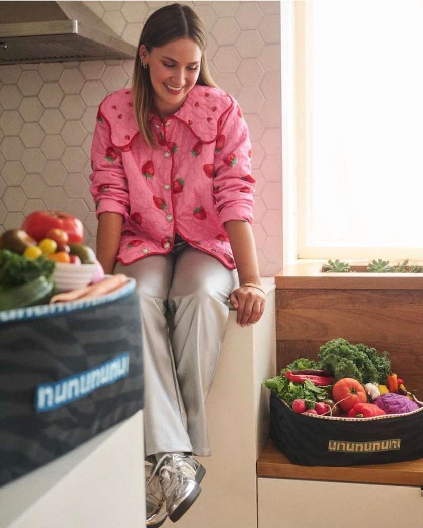 Woman in pink strawberry-print top sits on a counter with containers of fresh fruits and vegetables, enjoying a healthy and colourful meal.