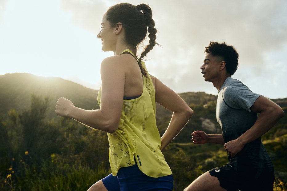 Woman in athletic wear running outdoors with a man, showcasing fitness and healthy lifestyle against a natural green landscape.