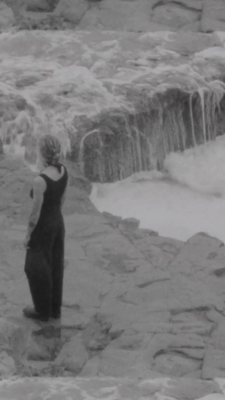 Woman in dark outfit standing on rocks looking at a small waterfall in black and white, captured in a natural outdoor setting.