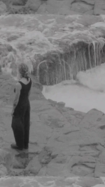Woman in dark outfit standing on rocks looking at a small waterfall in black and white, captured in a natural outdoor setting.