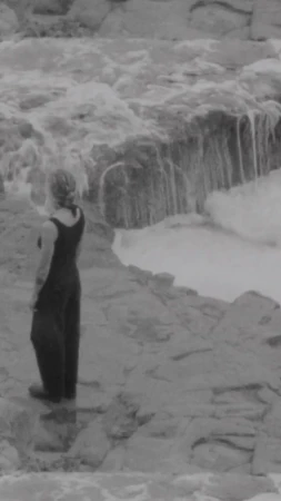 Woman in dark outfit standing on rocks looking at a small waterfall in black and white, captured in a natural outdoor setting.