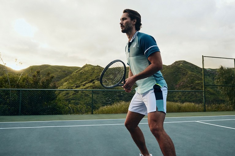 A man in tennis apparel holds a racquet on a tennis court in an outdoor setting with mountains in the background, sporty and athletic.