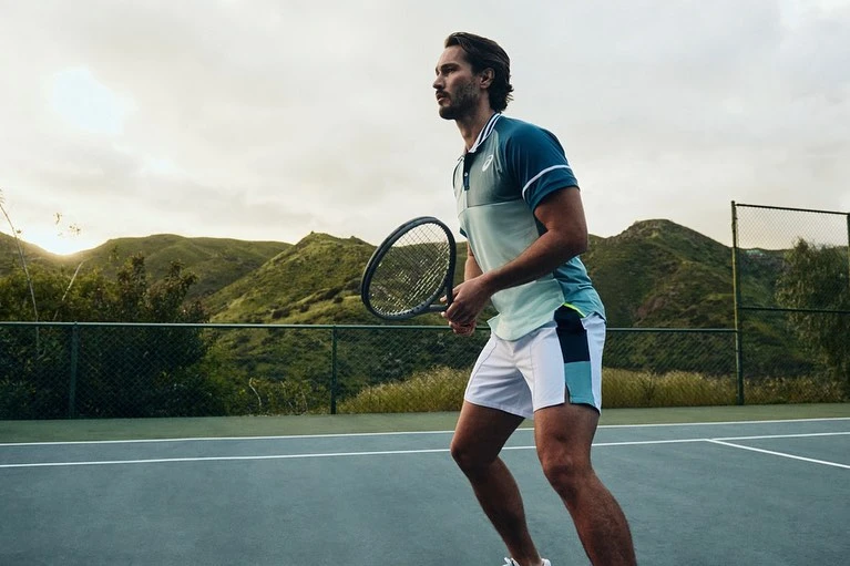 A man in tennis apparel holds a racquet on a tennis court in an outdoor setting with mountains in the background, sporty and athletic.