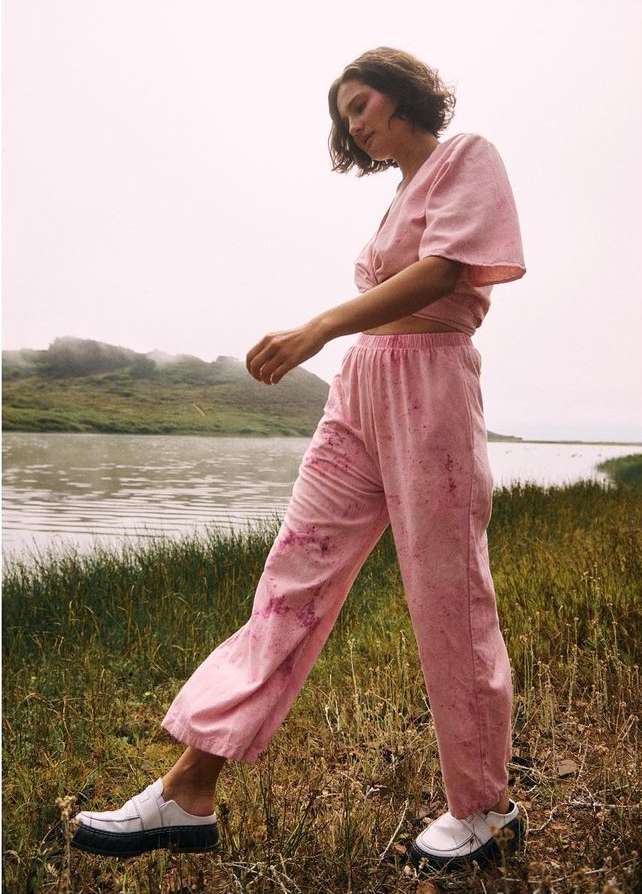 Woman in pink tie-front crop top and pink pants walking in field near lake on an overcast day, fashion editorial shot outdoors.