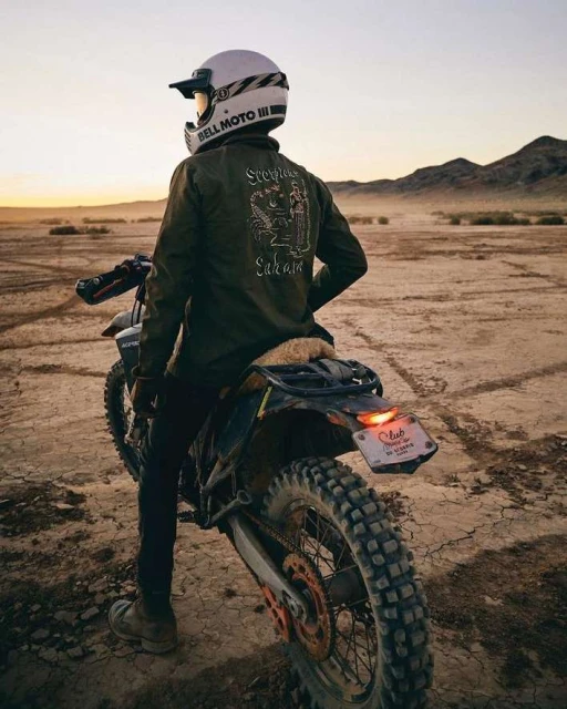 Adult in helmet and jacket sitting on a dirt bike in a desert landscape during sunset with mountains in the distance.