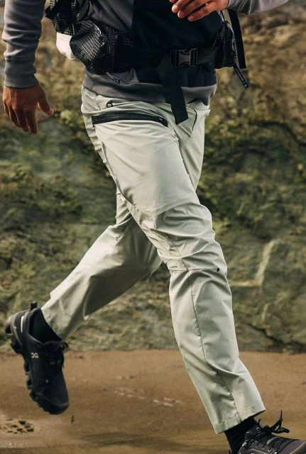Close-up of an adult wearing grey pants and black athletic shoes running on sand in front of a rocky outcrop, focus on lower body.