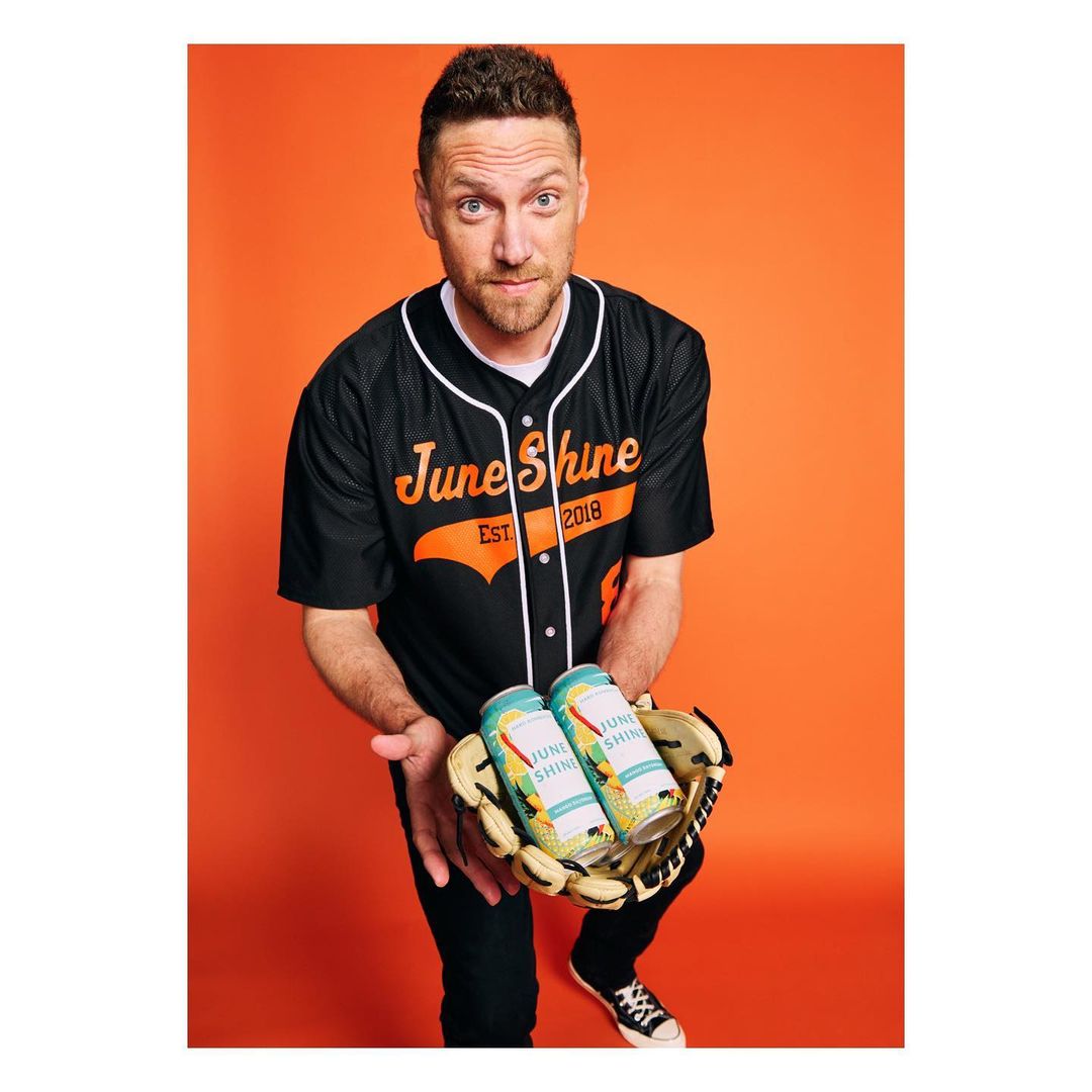 Adult male holds a baseball glove with two June Shine cans, wearing a baseball jersey against an orange backdrop, looking directly at the viewer.
