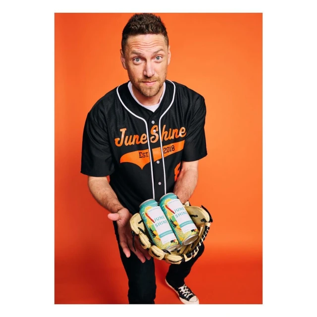 Adult male holds a baseball glove with two June Shine cans, wearing a baseball jersey against an orange backdrop, looking directly at the viewer.