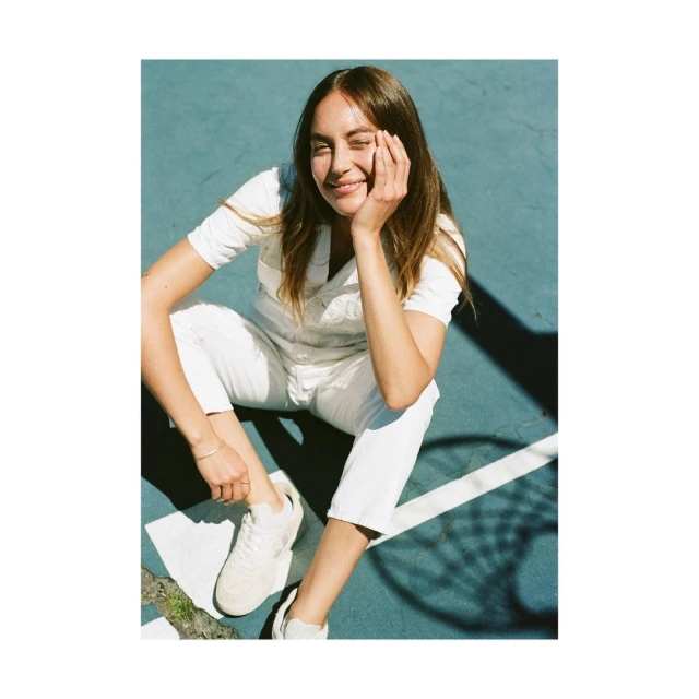 A young adult woman in a white jumpsuit sits smiling on a teal sports court with dark shadows, wearing white sneakers and a gold bracelet.