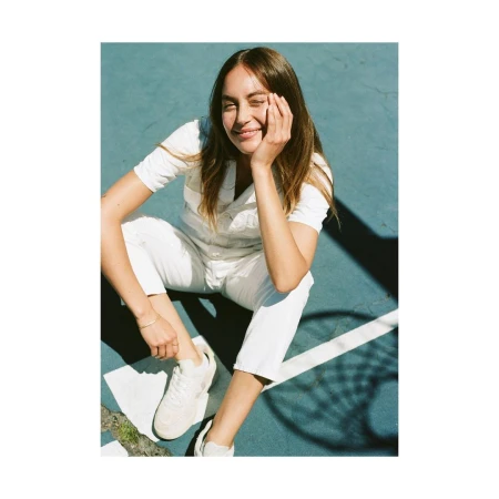 A young adult woman in a white jumpsuit sits smiling on a teal sports court with dark shadows, wearing white sneakers and a gold bracelet.