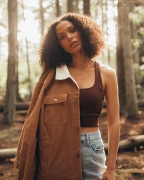 Woman with curly brown hair posing in a forest wearing a brown jacket draped over her shoulder, a tank top, and light jeans.