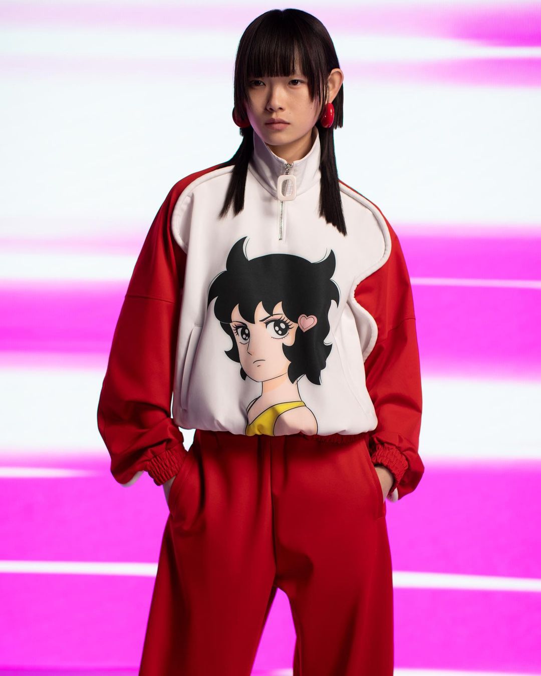 Asian woman in red track suit with anime graphic top poses in studio with pink and white background stripes, portrait framing.