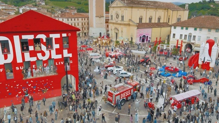 Aerial view of a bustling town square featuring a Diesel advertisement on a building, surrounded by a large crowd and various vehicles.