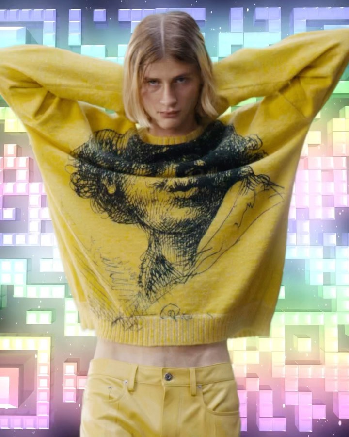Fashion shot of a person wearing a yellow graphic sweater and pants, arms raised, against a multicolored abstract background.