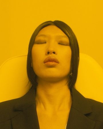 Close-up of an Asian adult woman with closed eyes and a black blazer, under yellow lighting. Simple background, focus on her face.