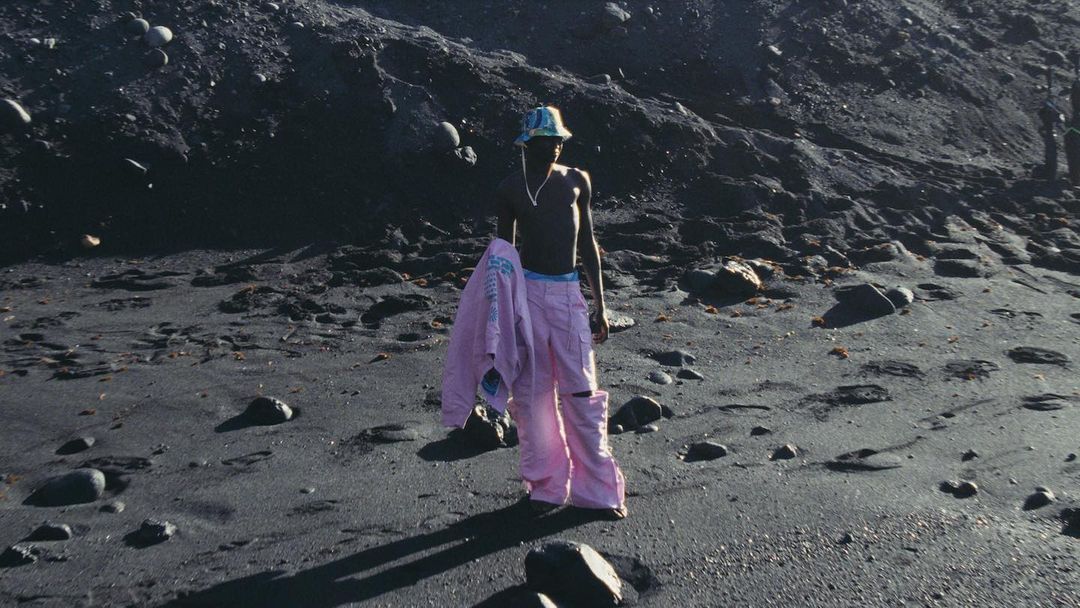 A shirtless man in pink pants and a bucket hat stands on a black sand beach with a jacket over his arm against a dark background.