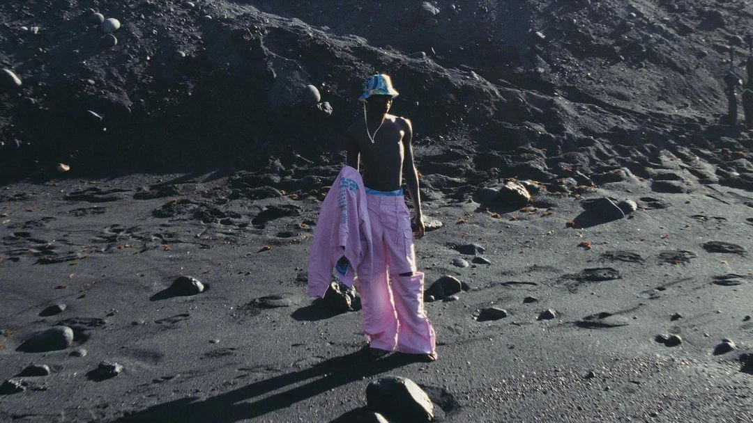 A shirtless man in pink pants and a bucket hat stands on a black sand beach with a jacket over his arm against a dark background.