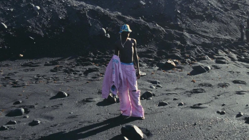 A shirtless man in pink pants and a bucket hat stands on a black sand beach with a jacket over his arm against a dark background.