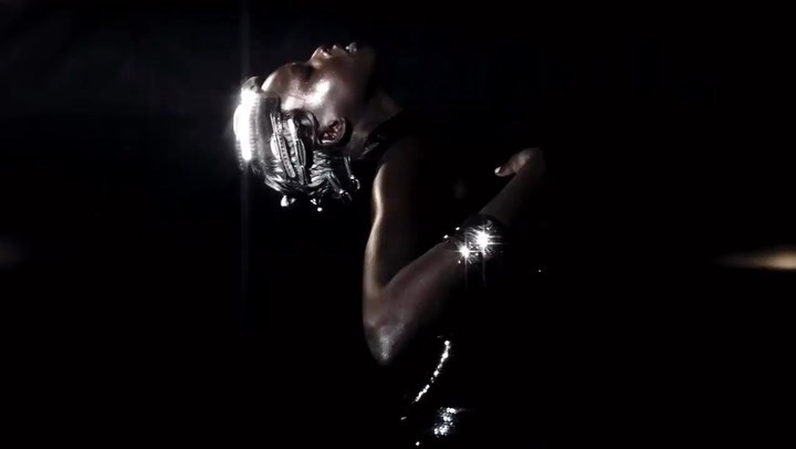 A human with silver body paint and a futuristic headset stands in the darkness, illuminated by soft light.