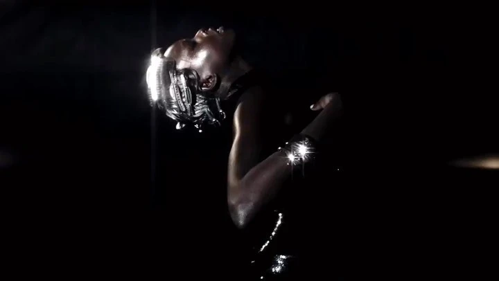 A human with silver body paint and a futuristic headset stands in the darkness, illuminated by soft light.