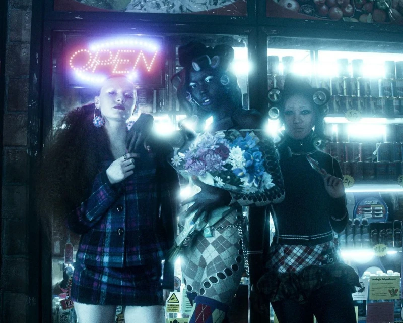 Three stylized women pose in front of a store with an OPEN neon sign, showcasing unique makeup, hair accessories, and diverse fashion choices.