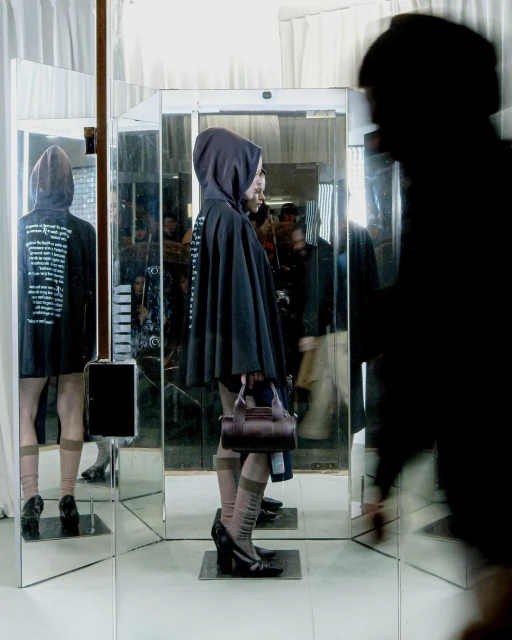 A woman in a hooded cloak is reflected in a series of mirrors while holding a brown handbag in an indoor setting.