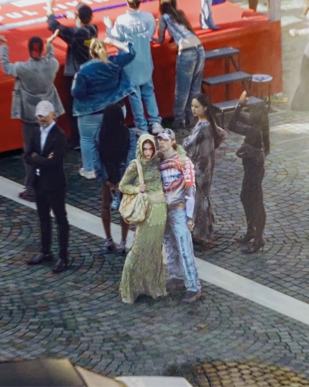 Fashionable group poses on cobblestone street with red barrier and onlookers in background. Street style, outdoor setting. 