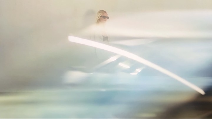 Blonde woman in sunglasses standing behind a blurred foreground element captured by Robbie Mailer-Howat